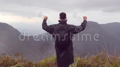 年轻的男徒步旅行者穿着雨衣，背包伸向<strong>山顶</strong>，举起双手。 男子游客<strong>站在</strong>